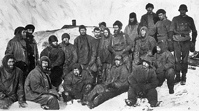 Группа мужчин в тяжелой зимней одежде и шляпах сидят тесно друг от друга. Их окружает снег и лед.
