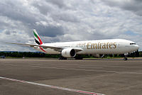 A mostly white Boeing 777, with some red, green and black markings, of Emirates. It is on a runway under a cloudy sky, facing right.