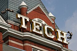 Large, white, capital letters spelling "TECH" situated  just below the pointed roof of the square, red brick tower of the  administration building