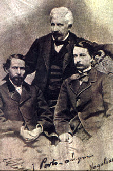 Старая фотография, на которой изображены два темноволосых мужчины, сидящие на переднем плане, и седой мужчина, стоящий позади.