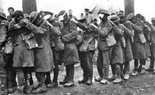 A column of British soldiers, most are holding the shoulder of the man in front of them. Many have bandages covering their eyes, others are covering their eyes with their hands.