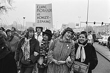 A black and white image of demonstrators marching with linked arms and carrying signs in support of Eliane Morissens' hunger strike.