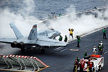 Chasseur F/A-18 sur le pont d'envol, avec l'équipage en train de le guider pour son catapultage.