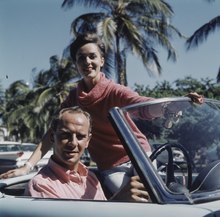 Photographie en couleur montrant Hugo Koblet au volant d'une voiture, sa compagne assise à ses côtés, tous deux vêtus d'un pull rose.