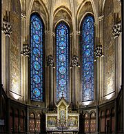 Photographie en couleurs de la chapelle vue de face avec au centre trois vitraux, de part et d'autre une mosaïque murale et en bas le retable.