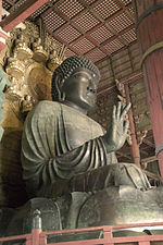 Three-quarter view and worm's-eye view of a seated statue. The palm of his right hand faces forward. The folds in his clothing are deeply sculpted. Behind the head of the sculpture there is a halo decorated with sitting statues. Color picture.