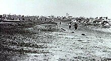 In the distance a group of people walk down a dirt road away from the village. The foreground shows vacant ground.