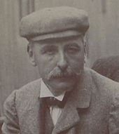 Photo en portrait d'un homme arborant une moustache en brosse et un couvre-chef.