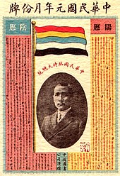 A calendar with a picture of a Chinese man in the centre. On top of it stands a flag with five horizontal stripes (red, yellow, blue, white, and black).