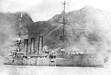 A large ship at anchor close to the shoreline, tall mountains directly behind the ship