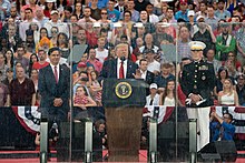 President Trump stands behind a plexiglass shield addressing the crowd. He is flanked by then-Acting Secretary of Defense Mark Esper on the left and Chairman of the Joint Chiefs of Staff Joseph Dunford on the right. A crowd sits on bleachers behind them.