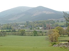 Photographie d'un paysage rural dominé par une haute colline ; au centre, les ruines d'une abbaye dans un cimetière.