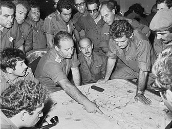 Réunion du Commandement régional du Nord de l'Armée de défense d'Israël lors de la guerre du Kippour, le 10 octobre 1973. L'homme qui pointe l'index est Yitzhak Hofi, celui aux deux mains sur la carte est David Elazar, chef d'État-Major, l'homme aux lunettes foncées se tenant debout derrière eux est Rehavam Zeevi. (définition réelle 1 800 × 1 350)