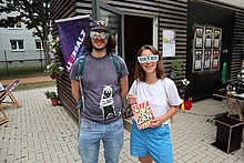 Ein Mann und eine Frau stehen vor einem Holzcontainer in einem Hof. Beide haben übergroße Papierbrillen auf. Sie hat eine Tüte Popcorn in der Hand.