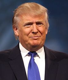 Trump at lectern before backdrop with elements of logo "TRUMP DonaldJTrump.com"