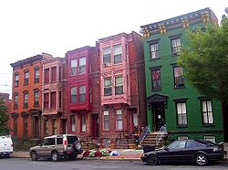 Группа из пяти трехэтажных кирпичных домов шириной в два и три бухты на городской улице. Один справа окрашен в зеленый цвет, три в центре имеют выступающие ниши на верхних этажах, а крайний левый неокрашен.