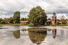 Un lac avec quelques nénuphars. De beaux arbres sur la berge, sous lesquels quelques bâtiments rouges se blottissent. Une petite tour donne l'heure.