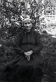 A photograph of a woman with a patterned dress and her hands in her lap sitting in front of a bush