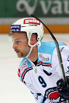 Photographie couleur d’un homme, la tête de profil, regardant vers la gauche, vêtu d’un maillot blanc aux épaules turquoise et d’un casque de hockey sur glace