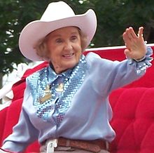 An elderly woman wearing a cowboy hat and blue shirt waves to unseen spectators.