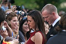A brown hair woman signs autographs for fans. She wears a red dress. Behind her there is a blond man dressed with a suit. The woman and the man are facing a crowd of fans.