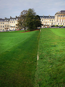 A ditch or trench in the grass with the right hand wall vertical and the left hand wall sloping. In the background are trees and yellow coloured stone buildings.