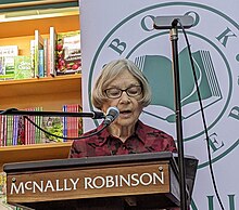Sarah Klassen is shown reading from The Russian Daughter at the book's launch at McNally Robinson Boookstore, Winnipeg in 2023. Klassen has short grey hair, glasses, and light coloured skin. They are wearing a collared shirt in a dark red print. They are behind a wooden podium with the words McNally Robinson printed on it. The store's bookshelves are visible behind the reader. So is a banner with the bookstore's logo.