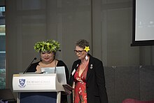 Two women in dresses look at their notes at a lecturn