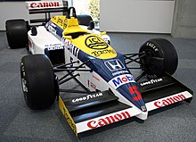Photographie en couleur d'une Formule 1 bleu foncé, blanche et jaune, vue de trois-quarts droite, exposée dans un musée.