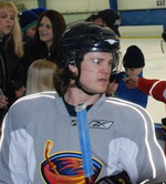 A Caucasian hockey player shown from the chest up. He wears a gray jersey with a stylized brown thrasher holding a hockey stick as a logo and a black helmet with a visor