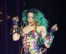 Laganga, on stage, glances down at the crowd. She is seen from the waste up, wearing a teal wig and a bikini top attached to a lattice of multi-colored straps. This garment is matched with gauntlet-like gloves.