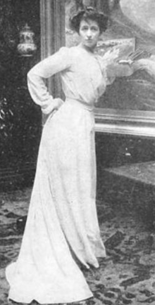 A white woman wearing a long white skirt, standing in front of a large painting