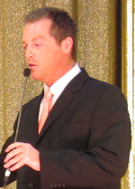 A man with dark hair wearing, including a black suite with a pink tie.