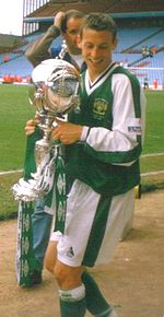 A brown-haired footballer holding a silver trophy adorned with green ribbons. He is wearing a green jersey with white sleeves, and white shorts.