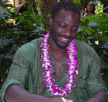A black man, wearing a green shirt and a necklace of purple flowers.