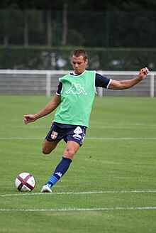 Photographie montrant un footballeur qui porte un survêtement bleu et un chasuble vert s'apprêtant à tirer dans un ballon de football