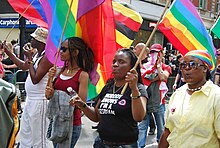 Photographie de plusieurs femmes noires à la Pride tenant des drapeaux arc-en-ciel. Une porte un t-shirt «Nobody knows I'm a lesbian ».