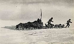 A loaded sledge being pulled across an icy surface by two figures and a team of dogs