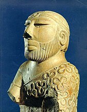 A carved stone statue of a bearded man with a prominent nose wearing a garment with a pattern