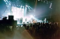 Рок-группа Oasis выступает на сцене перед большим проекционным экраном с изображениями на нем. У четырех участников привязанные к ним гитары.