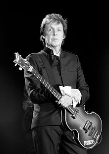 Black and white photgraph of McCartney standing onstage holding a bass guitar. He is wearing a dark suit.