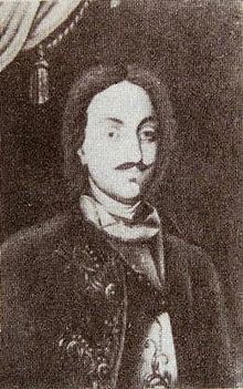 Portrait en buste en noir et blanc d'un homme mince aux cheveux mi-longs et à fine moustache portant une cravate de soie et une veste richement ornée