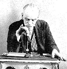 Photographie en noir et blanc d'un homme assis à une table et utilisant un pendule.