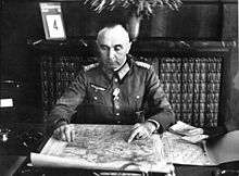 Photographie en noir et blanc d'un homme en uniforme assis sur son bureau et en train de scruter une carte.