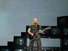 Мадонна в оливково-зеленой одежде поет в микрофон, держа в руках черную электрогитару.