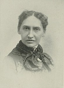 B&W portrait photo of a middle-aged woman with her hair in an up-do, wearing a dark, high-collared blouse.