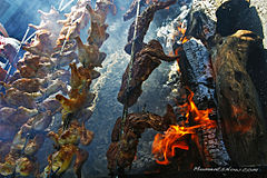Many whole chickens on large skewers over an open flame and a bed of coals.