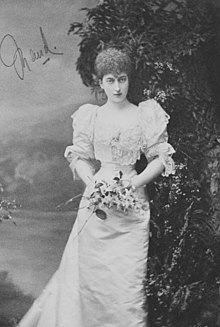 Old autographed black-and-white photograph of a young woman facing the camera, in a long white gown, with a very small waist and a bouquet of flowers.