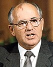 The image is a portrait of a middle-aged man (Mikhail Gorbachev) with short, light-colored hair, wearing large-framed glasses and a dark suit with a light-colored shirt and a dark tie. He has a noticeable red birthmark on his forehead. The background of the photo is indistinct.