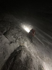 Bergretter klettert am Seil gesichert eine vereiste und verschneite Felswand bei Nacht im Schneesturm empor.